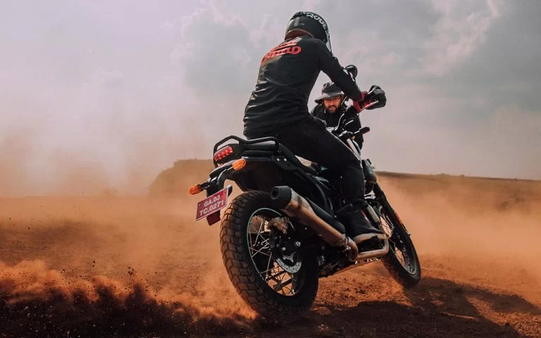 Actiefoto van achterkant genomen met motorrijder staand op de voetsteunen van een Royal Enfield Scram 411 en wegscheurt in een gravelachtig terrein