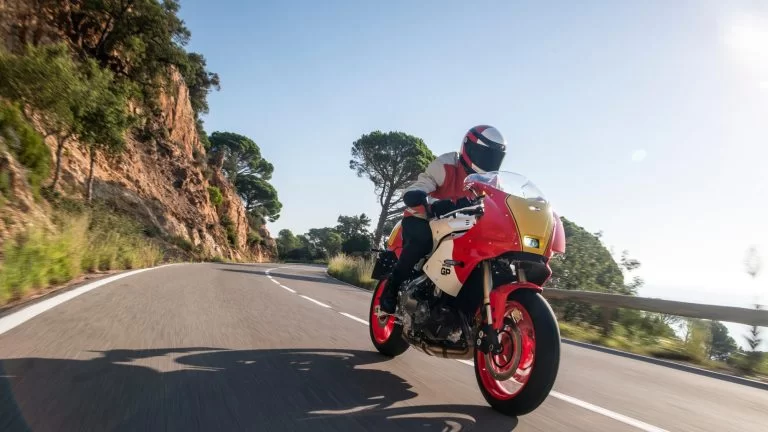 Motorrijder rijdt op een Heritage gp motor met rood en gele accenten langs een bergwand en water