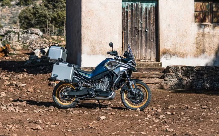 Lifestylefoto van CF Moto 800MT Touring blauw met koffers. Op achtergrond een oud vervallen berghuisje.