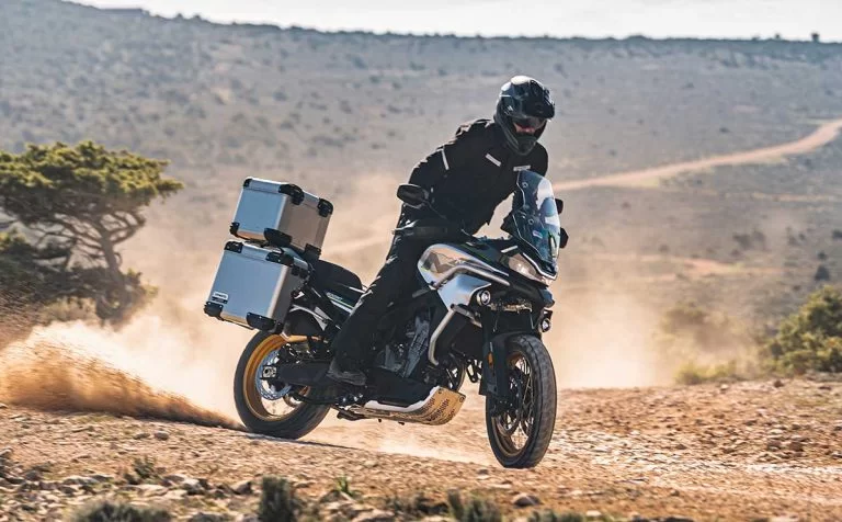 Actiefoto van CF Moto 800MT Touring blauw met koffers met motorrijder op bergachtig terrein en heuvels op achtergrond